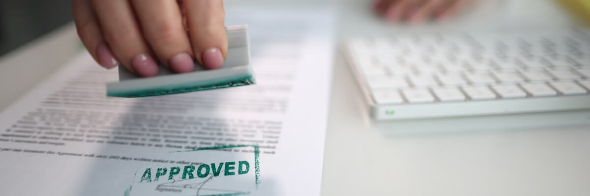 Eine Nahaufnahme zeigt eine Hand mit rosa lackierten Fingernägeln, die einen grünen Stempel mit der gut lesbaren Aufschrift „APPROVED“ auf ein Dokument drückt. Der Abdruck „APPROVED“ ist deutlich auf dem Papier zu sehen.